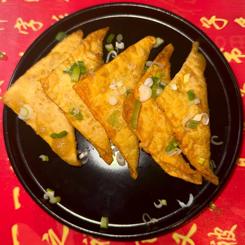 Golden-fried wontons, garnished with chopped green onions, served on a black plate over a red tablecloth.