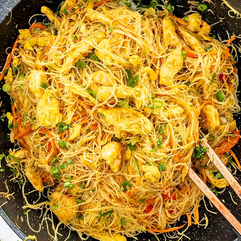 Close-up of Singapore noodles with chicken, shrimp, vegetables, and chopsticks in a black wok.