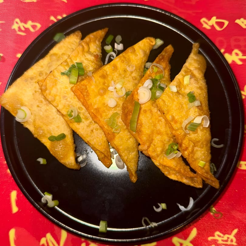 Golden-fried wontons, garnished with chopped green onions, arranged on a black plate over a red patterned tablecloth.