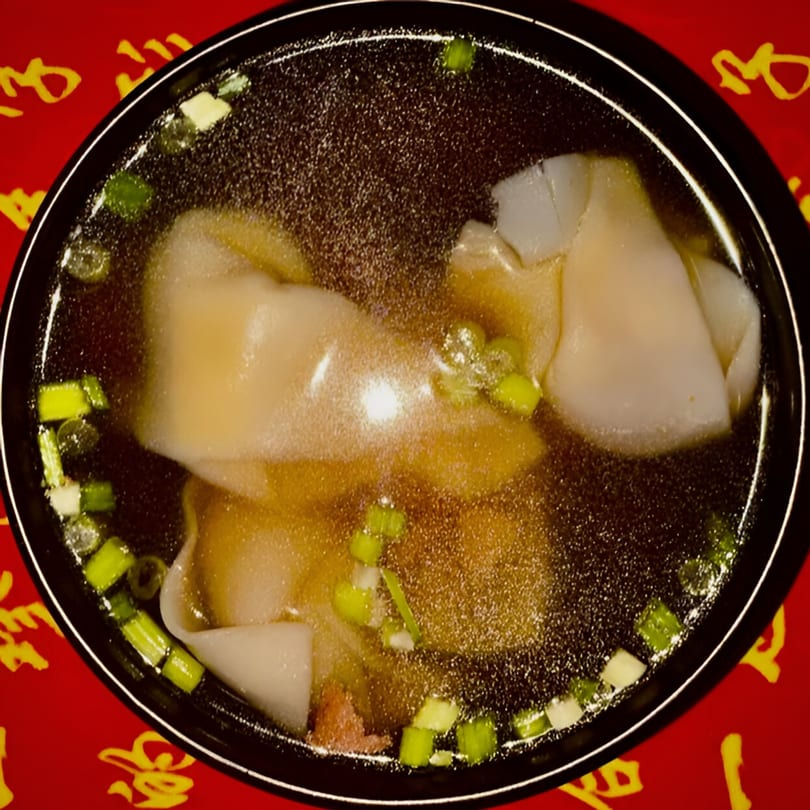 A close-up of a bowl of Chinese wonton soup with scallions on a red patterned surface.