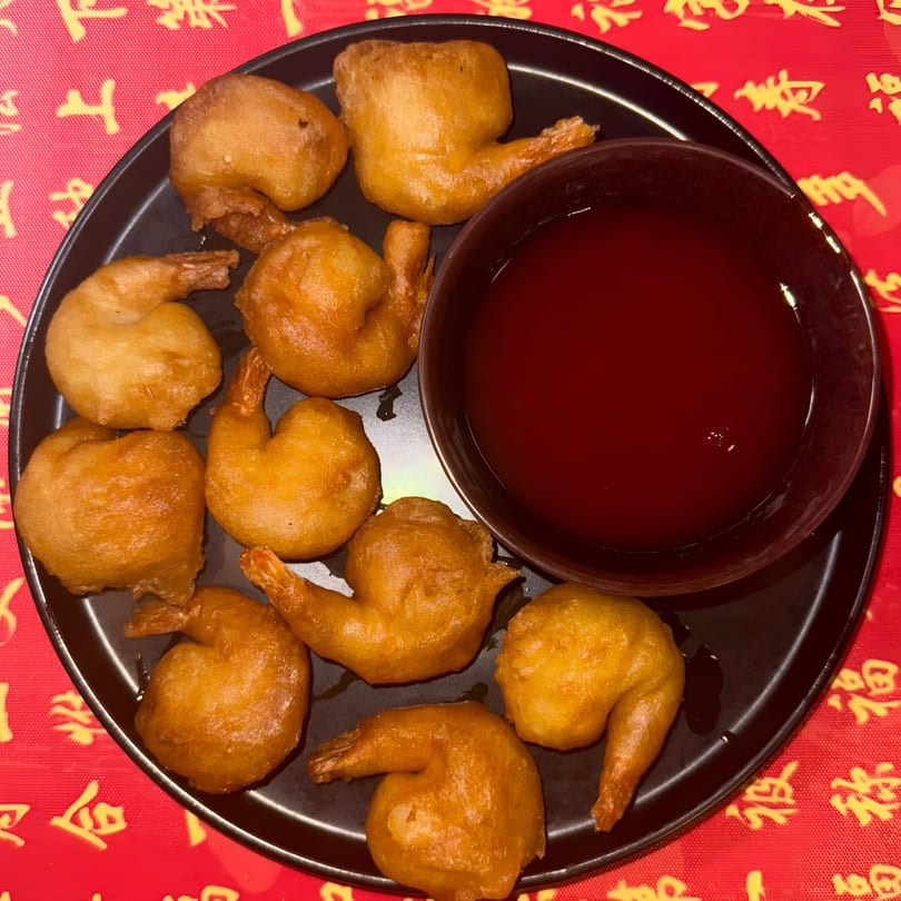 A black plate of crispy fried shrimp with a side of red sweet and sour sauce on a red tablecloth.