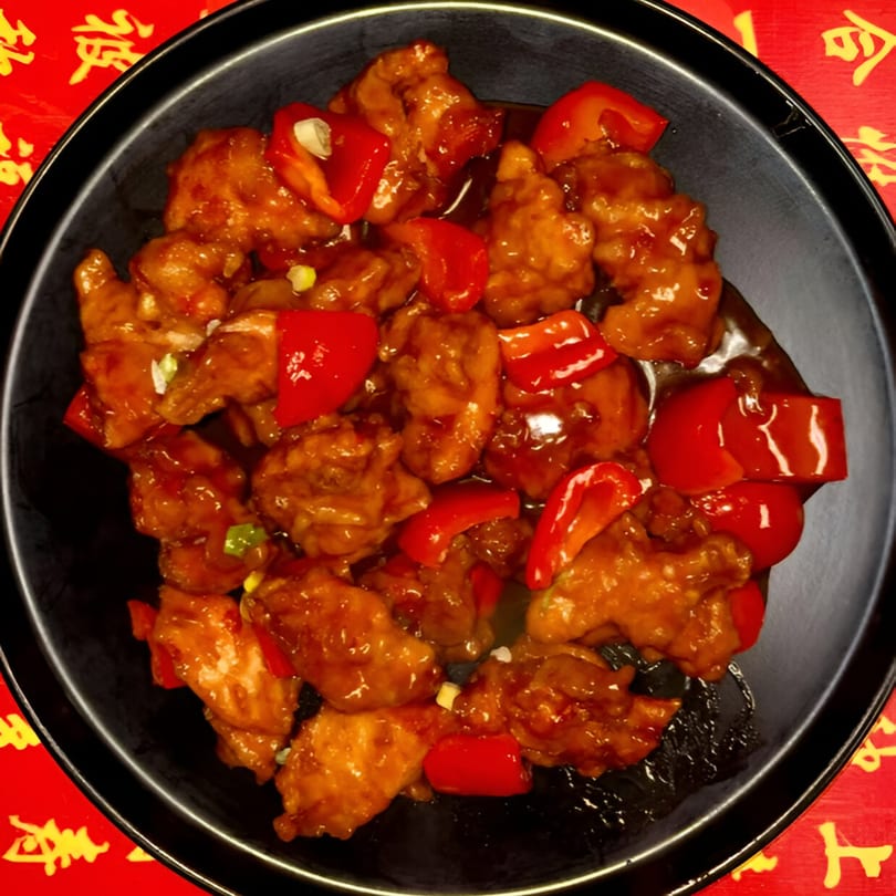 Sweet and sour chicken with red bell peppers on a black plate, against a red background.