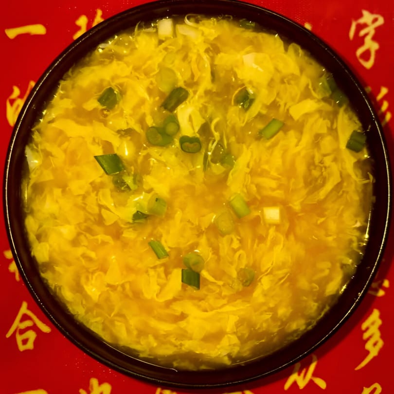 A bowl of Red Lantern's Chinese egg drop soup with green onions on a red background.