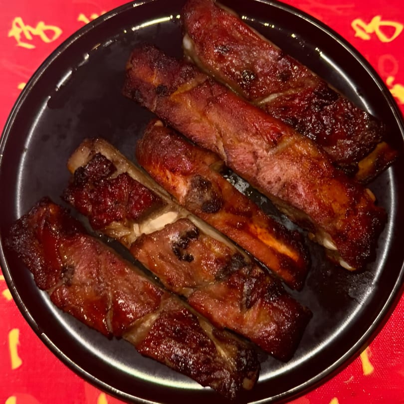 A black plate filled with several pieces of glistening, dark brown Chinese BBQ pork ribs.